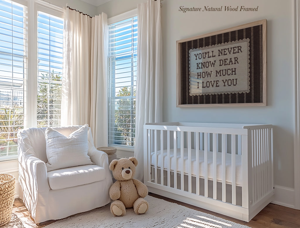 A cozy nursery with a white crib, a white chair, and a teddy bear. The room has large windows that let in natural light, and a framed sign on the wall that reads "You'll never know how much I love you".