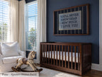 A nursery room with a wooden crib, a rocking horse, and a framed sign with a message. The room has blue walls and large windows that let in natural light.