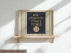 A framed poster with the text "ALL LEGENDS START SMALL" and a baseball graphic on it, displayed on a wooden shelf against a white wall.
