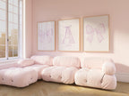 A cozy living room with a large window, featuring three framed pictures of bows on the wall. The room has a pink color scheme, with plush furniture and a wooden floor.