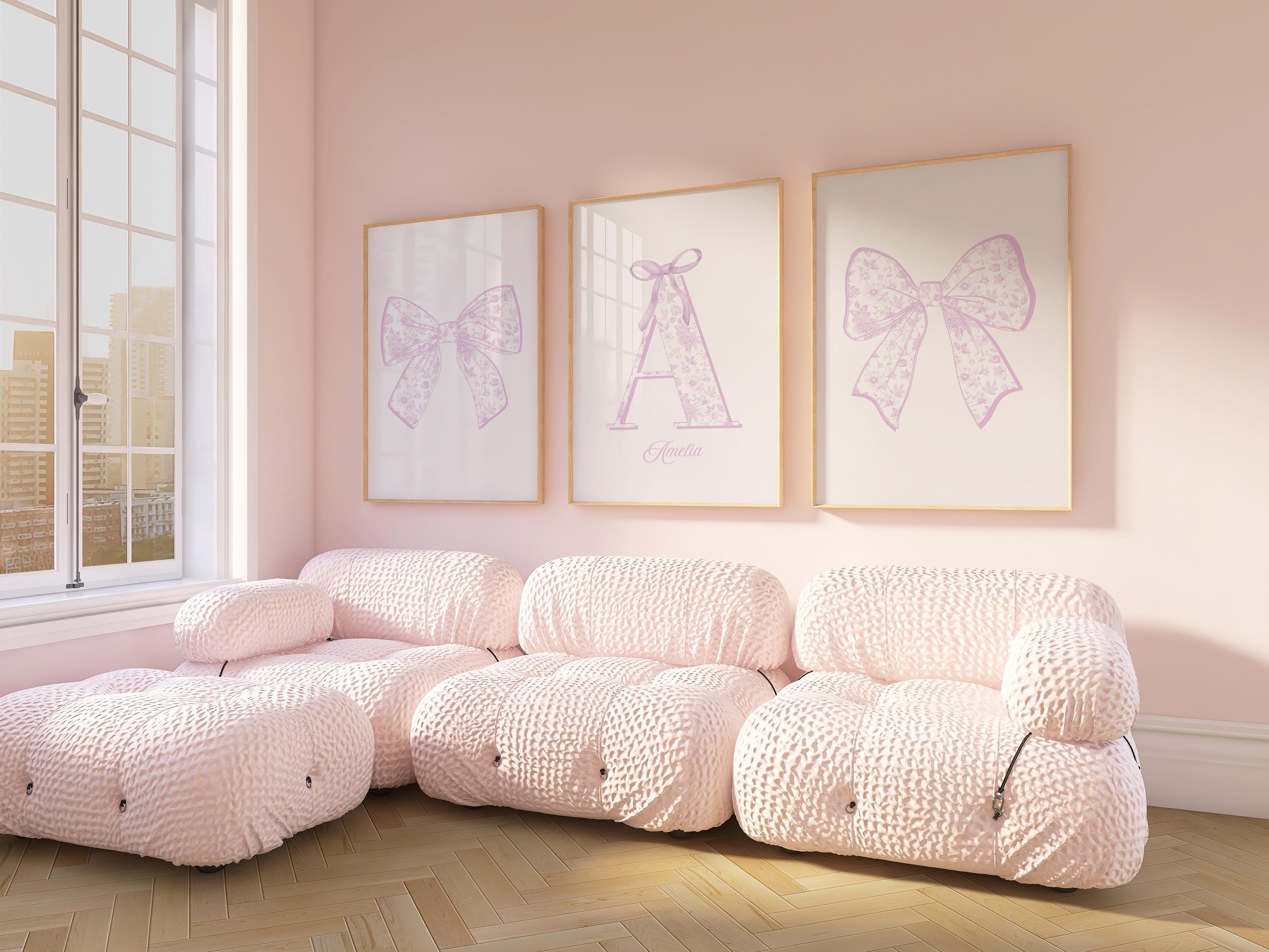 A cozy living room with a large window, featuring three framed pictures of bows on the wall. The room has a pink color scheme, with plush furniture and a wooden floor.