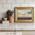 a framed landscape painting of a cloudy sky, placed on a marble countertop next to a potted plant and a stack of books.