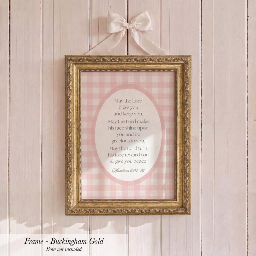 A framed print with a pink and white checkered background hangs on a wooden wall. The frame is gold and has a bow on top.