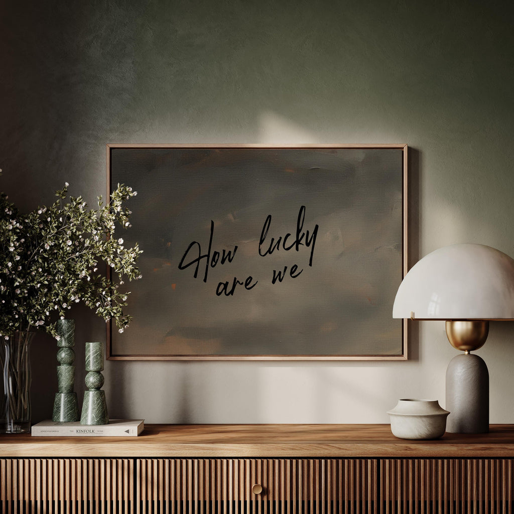 a wooden shelf with a framed artwork hanging on the wall above it, a lamp, and some decorative items such as a vase with flowers and a book.