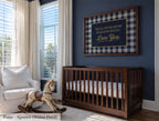 A nursery room with a wooden crib, a rocking horse, and a framed sign with a message. The room has blue walls and white curtains, creating a calm and serene atmosphere.