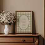A framed print with a quote about gift-giving is displayed on a wooden dresser next to a white vase filled with white flowers.