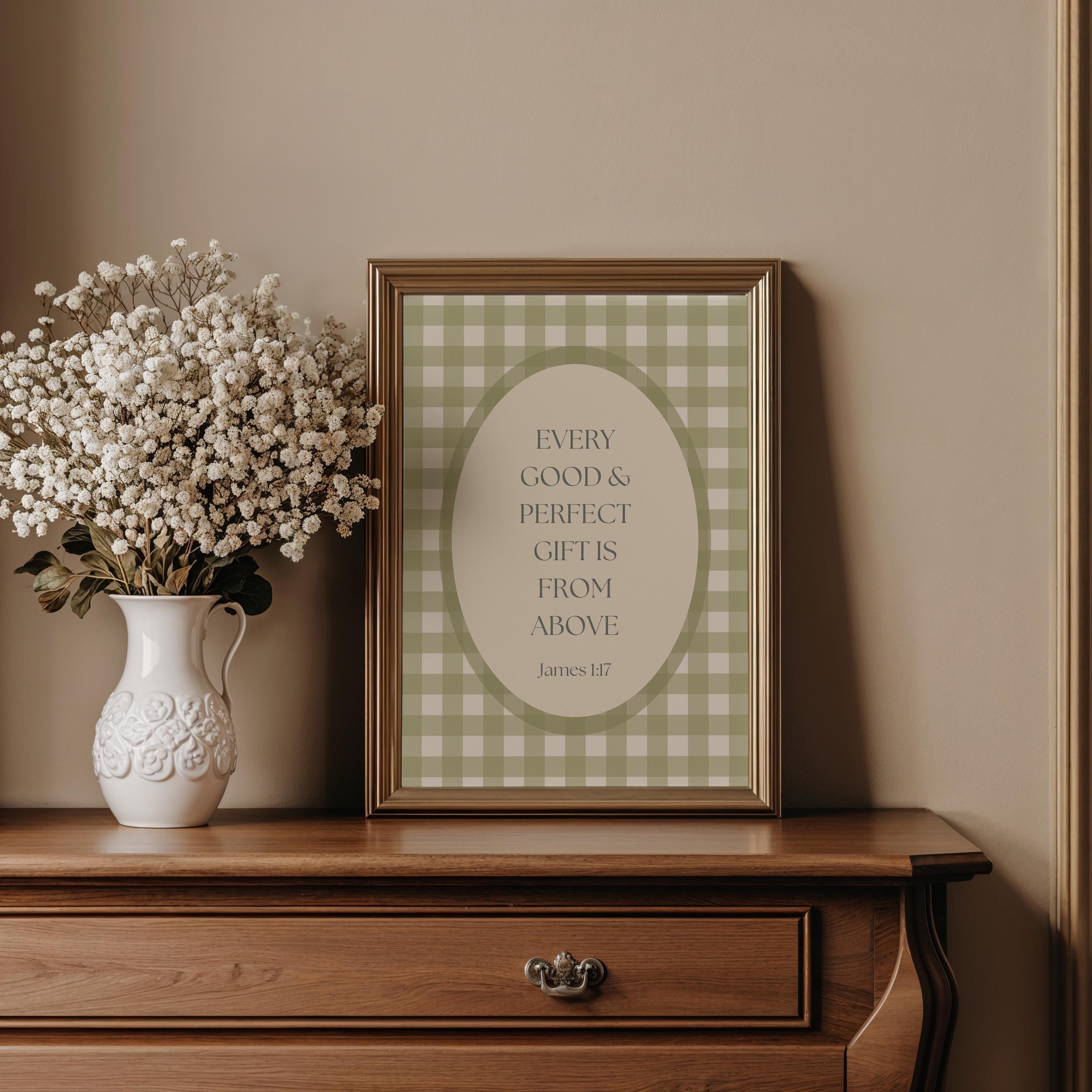 A framed print with a quote about gift-giving is displayed on a wooden dresser next to a white vase filled with white flowers.