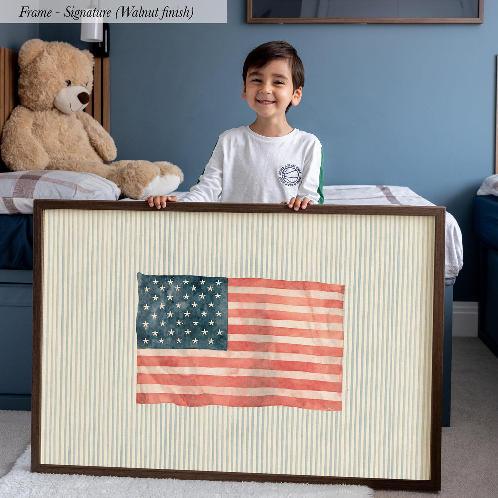 A young boy is standing in a bedroom, smiling and holding a framed picture of an American flag.