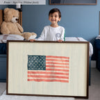 A young boy is standing in a bedroom, smiling and holding a framed picture of an American flag.