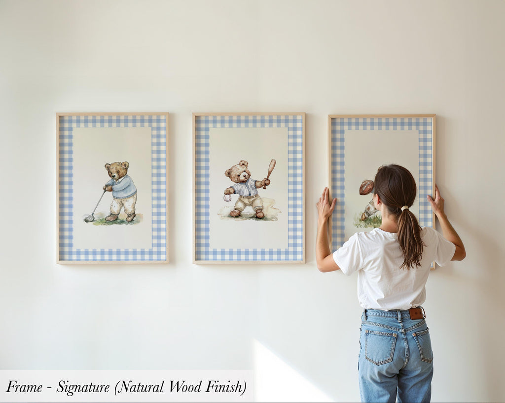A woman is hanging three framed pictures on a wall, each depicting a teddy bear in different poses.