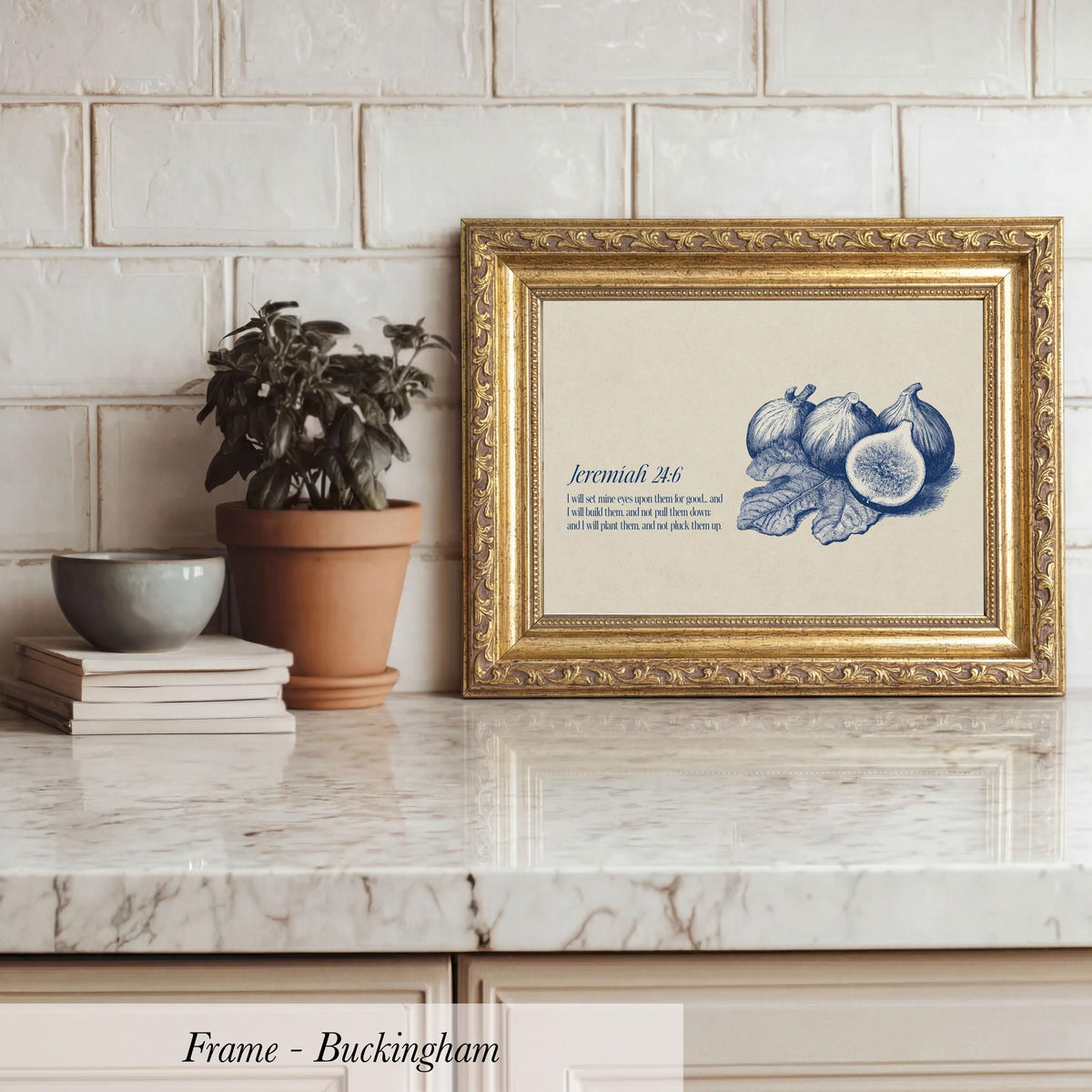 A framed print of a blue and white illustration of fruits, including figs and leaves, is displayed on a marble countertop next to a potted plant and a stack of books.