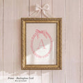 a gold-framed picture hanging on a wooden wall, with a pink bow tied around the top of the frame.