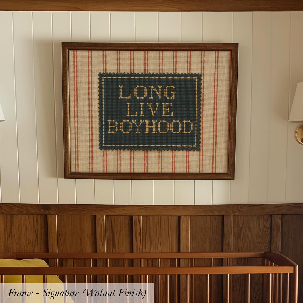 A framed cross-stitch design with the text "LONG LIVE BOYHOOD" on it, hanging on a wall above a wooden crib.