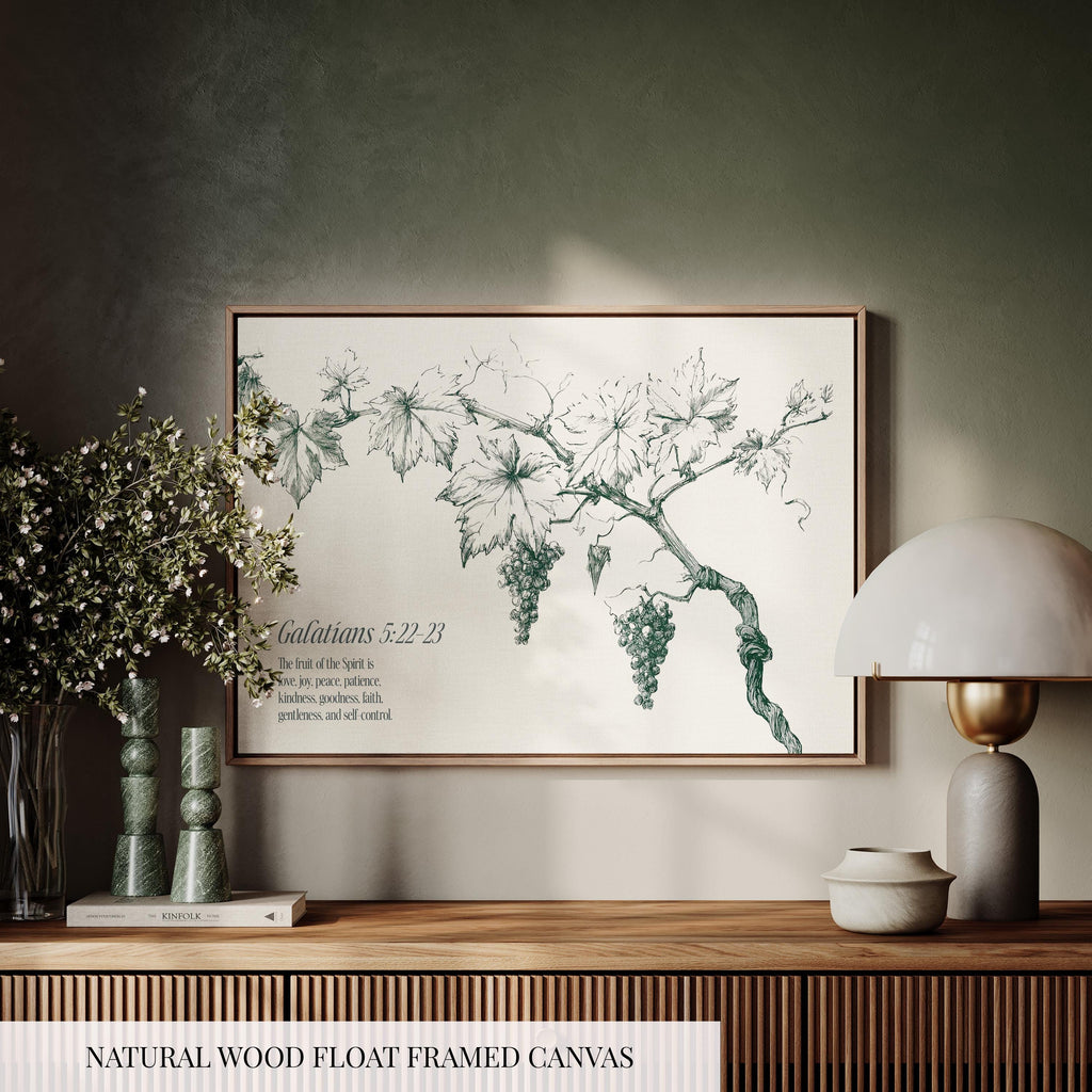 a framed artwork of a vine with grapes hanging from it, displayed on a wooden shelf. The artwork is surrounded by various decorative items, including a lamp, a vase with flowers, and a book. The text "Galatians 5:22-23" is visible, which is likely a biblical verse from the book of Galatians.