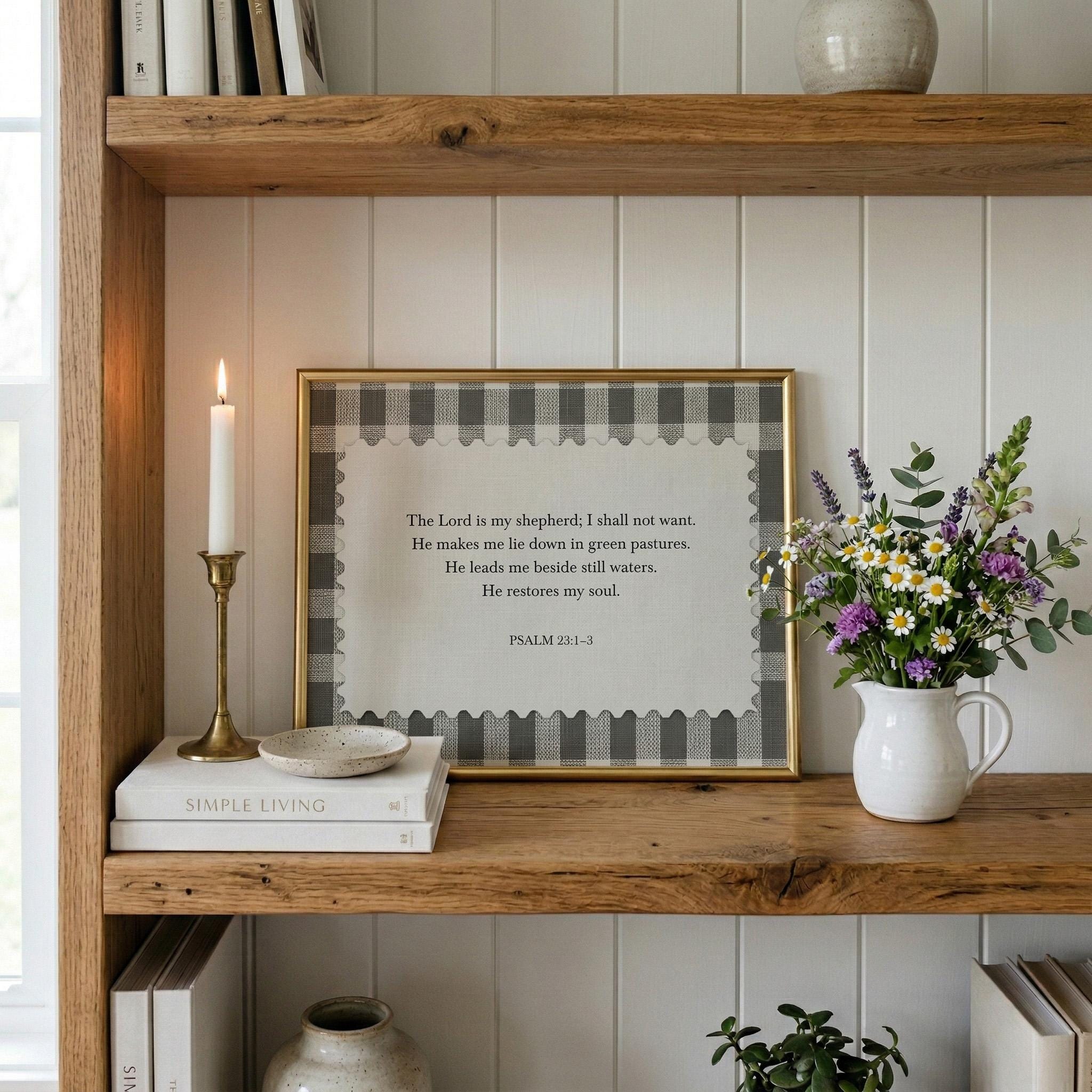 a wooden bookshelf with a framed Bible verse, a white candle, a white vase with purple flowers, and a stack of books.