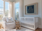 A cozy nursery with a white crib, a white chair, and a teddy bear. The room has large windows that let in natural light, and a framed sign with a message hangs on the wall.