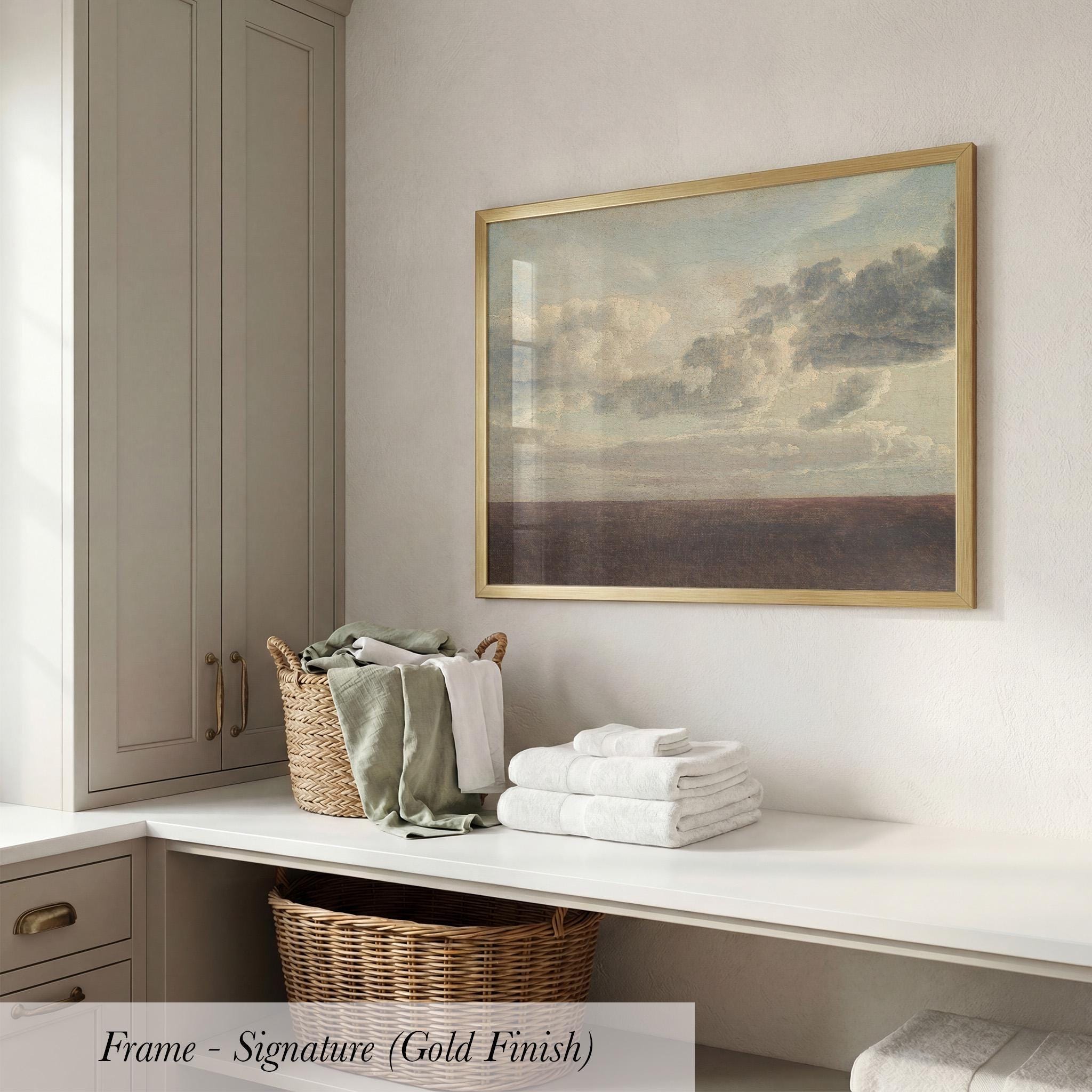 a framed painting of a cloudy sky hanging on a wall above a white countertop with folded towels and a wicker basket.