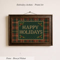 A framed embroidered design with the text "HAPPY HOLIDAYS" on a green plaid background, hanging on a wall.