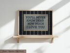 a framed cross-stitch design with the text "YOU'LL NEVER KNOW DEAR HOW MUCH I LOVE YOU" displayed on it.