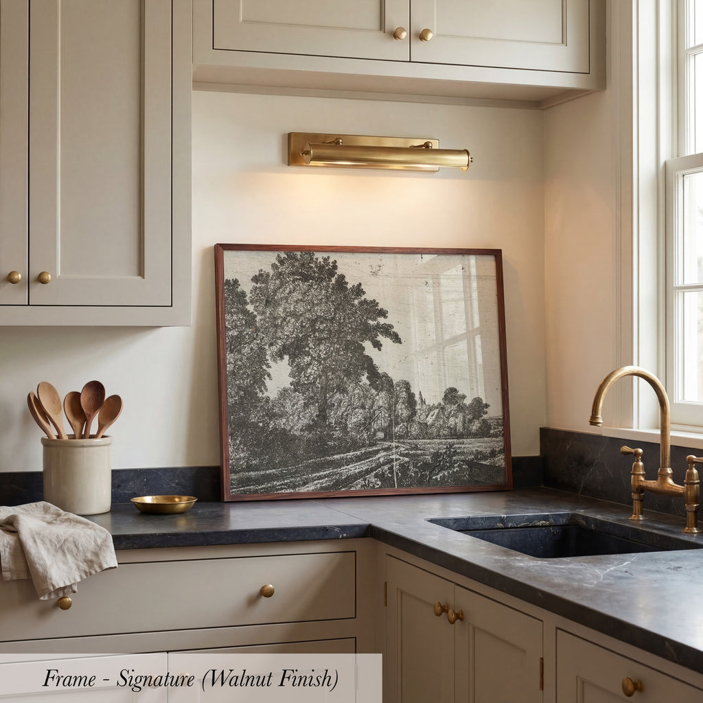 a kitchen with a framed black and white landscape painting on the wall above a sink. The painting depicts a serene scene of trees and a winding road. The kitchen has white cabinets and a marble countertop, creating a clean and elegant atmosphere.