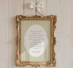 a gold-framed picture hanging on a white wall, displaying a green and white striped background with a white oval containing a poem or message.