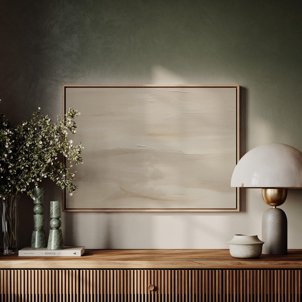 a wooden shelf with a framed painting, a lamp, and a vase of flowers. The painting is a large, blank canvas with a light beige background. The lamp has a white shade and a gold base, and the vase is filled with white flowers. The shelf also has a book and a small white bowl.