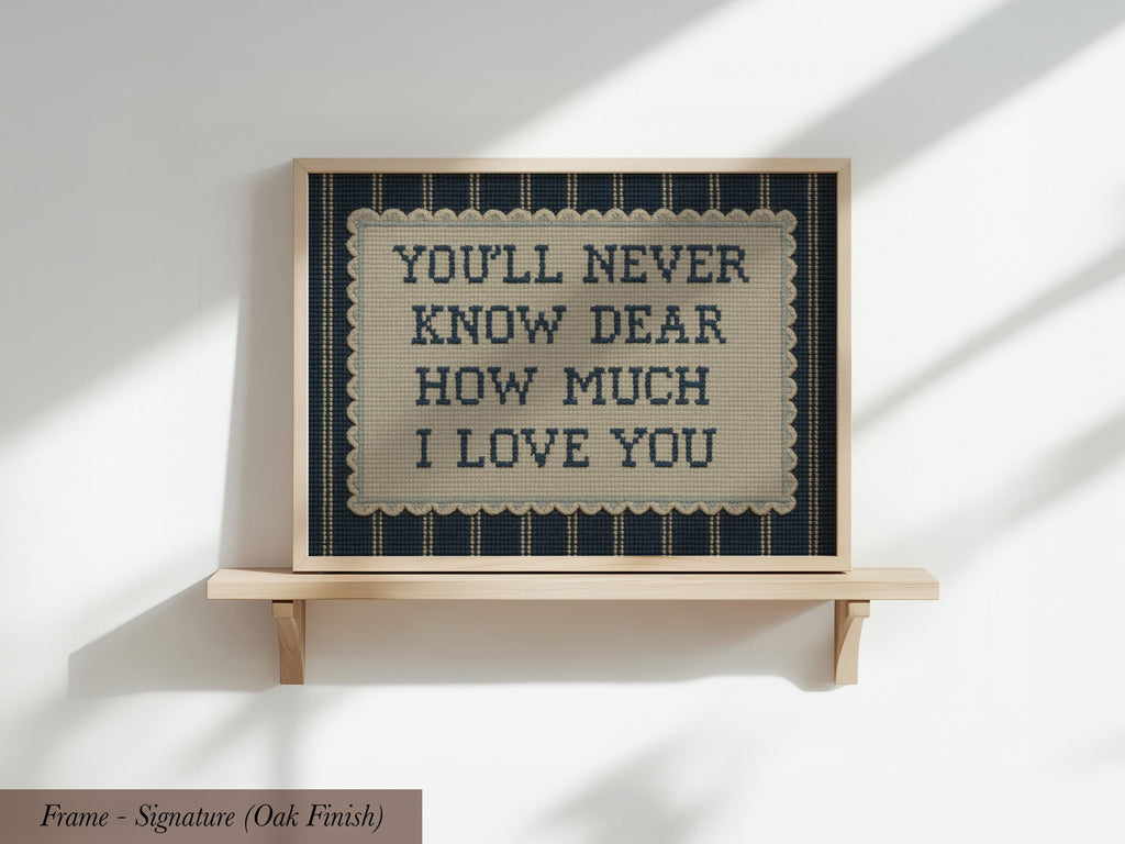 a framed sign with the text "YOU'LL NEVER KNOW DEAR HOW MUCH I LOVE YOU" on it, which is displayed on a wooden shelf against a white wall.