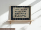 a framed sign with the text "YOU'LL NEVER KNOW DEAR HOW MUCH I LOVE YOU" on it, which is displayed on a wooden shelf against a white wall.