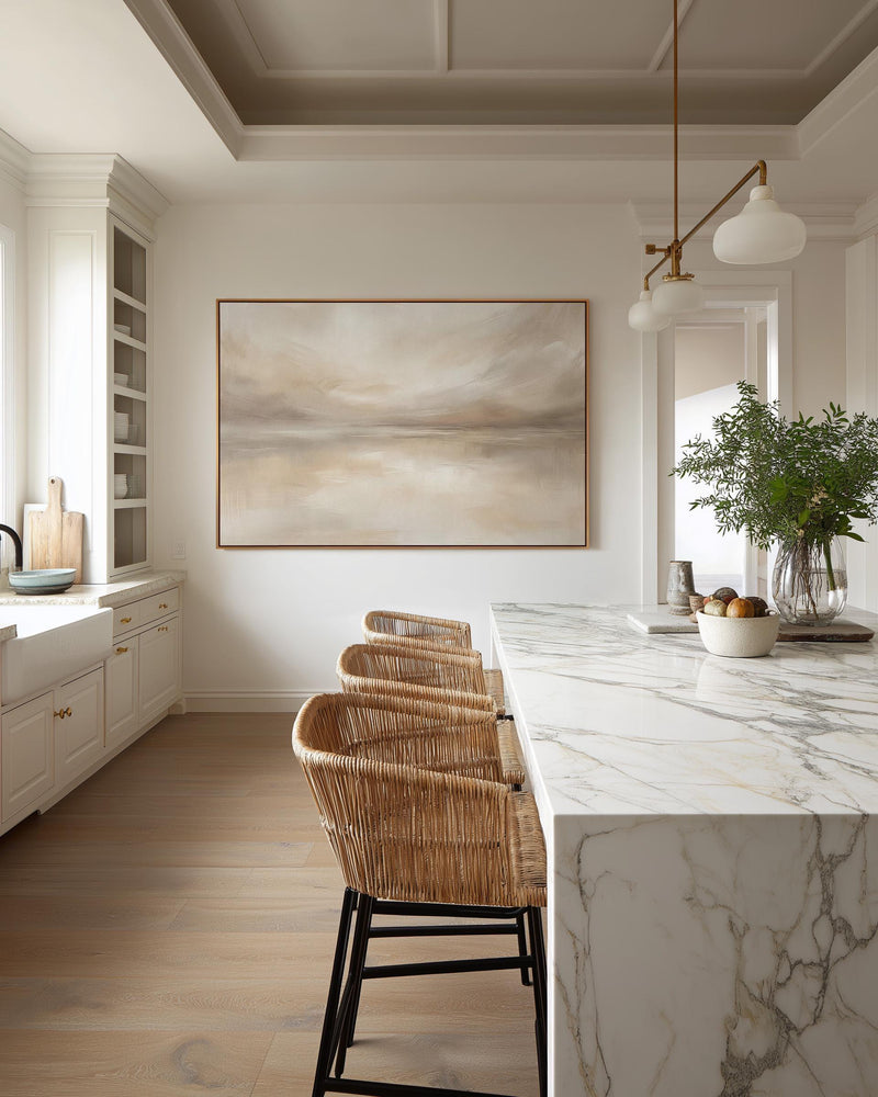 May include: A modern kitchen with a large abstract landscape painting in a gold frame. Three woven rattan bar stools with black metal legs sit in front of a marble countertop. A vase of greenery and fruit adds a natural touch.
