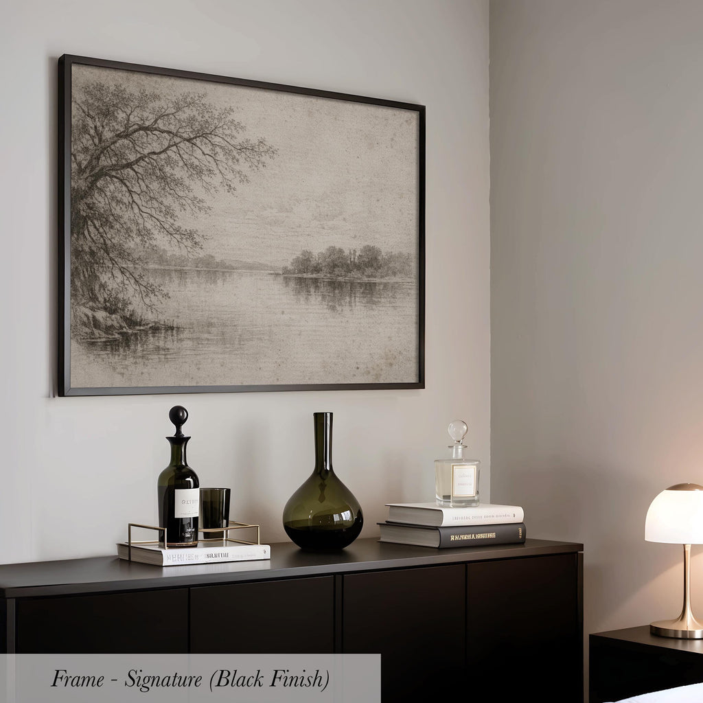 a black and white framed landscape painting hanging on a wall above a black cabinet with various decorative items on top, including a bottle, a vase, and a stack of books.