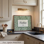 A framed artwork with a green floral pattern and the name "Beckham" is displayed on a kitchen counter, with a sink and cabinets in the background.