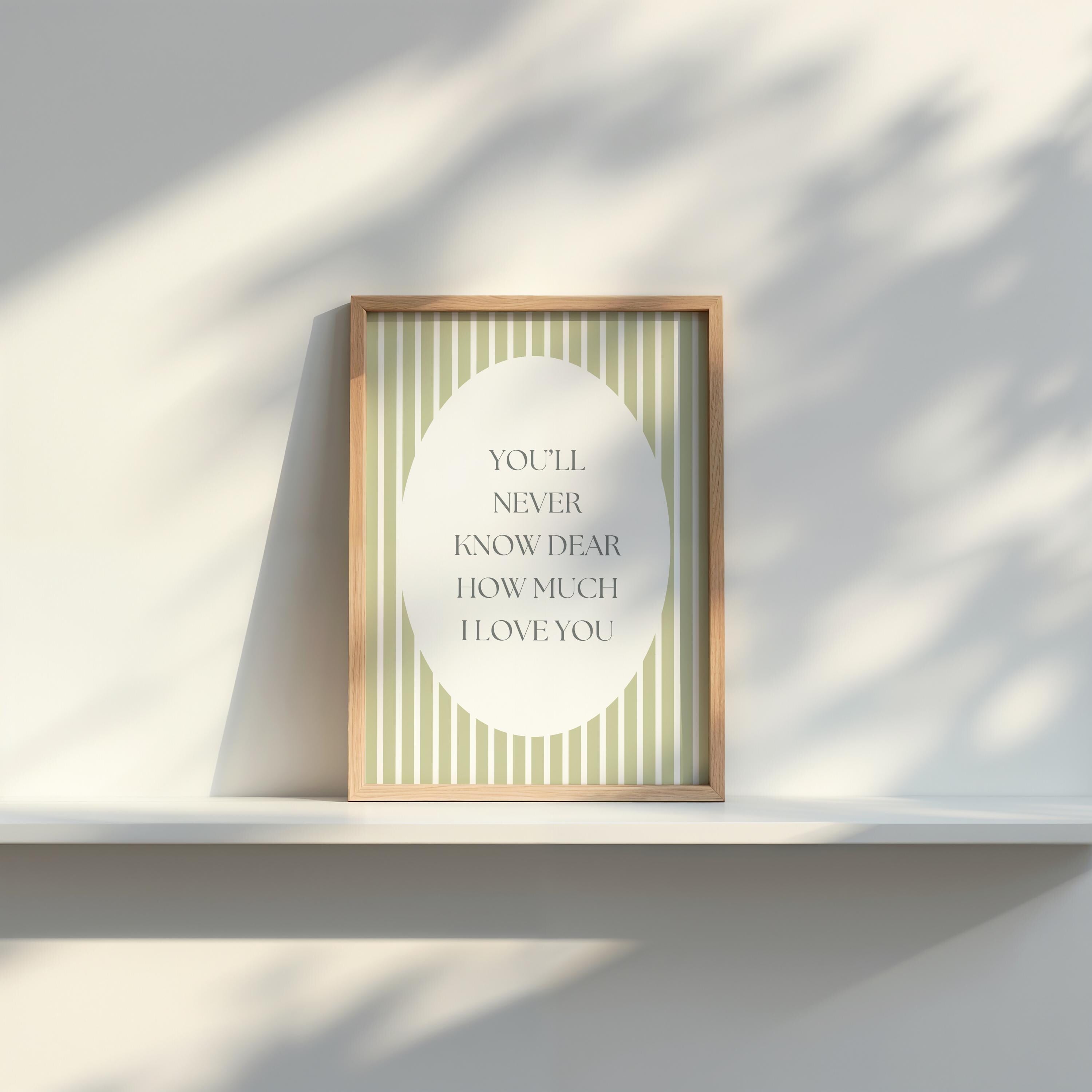 A framed poster with the text "YOU'LL NEVER KNOW DEAR HOW MUCH I LOVE YOU" is displayed on a white shelf against a white wall.