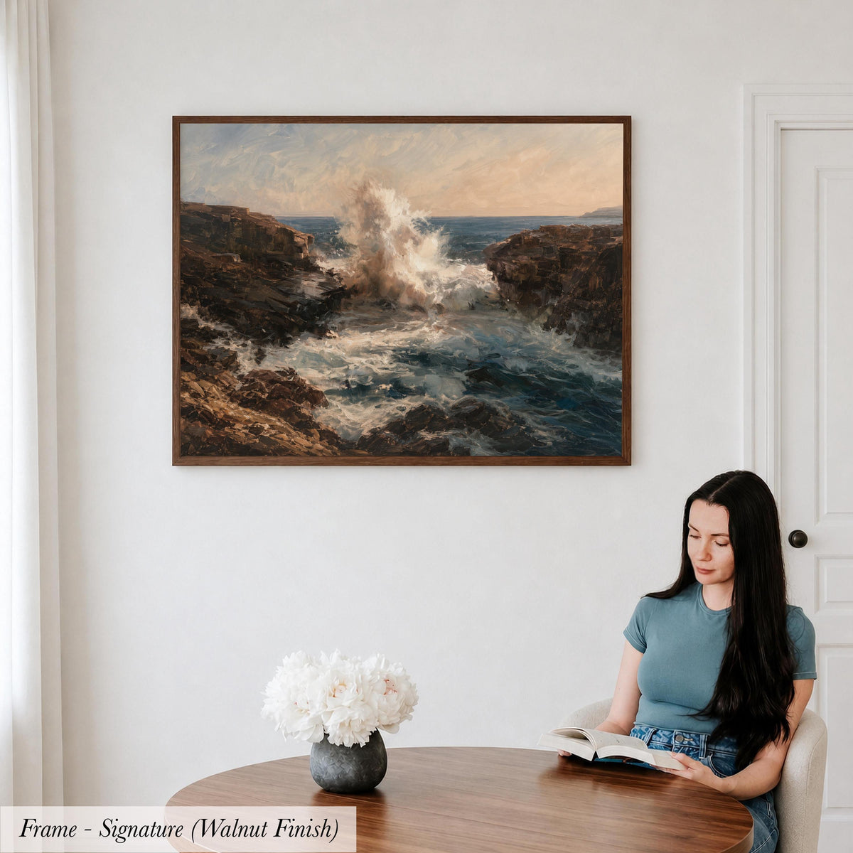 A woman is sitting at a table, reading a book, with a framed painting of a crashing ocean scene hanging on the wall behind her.