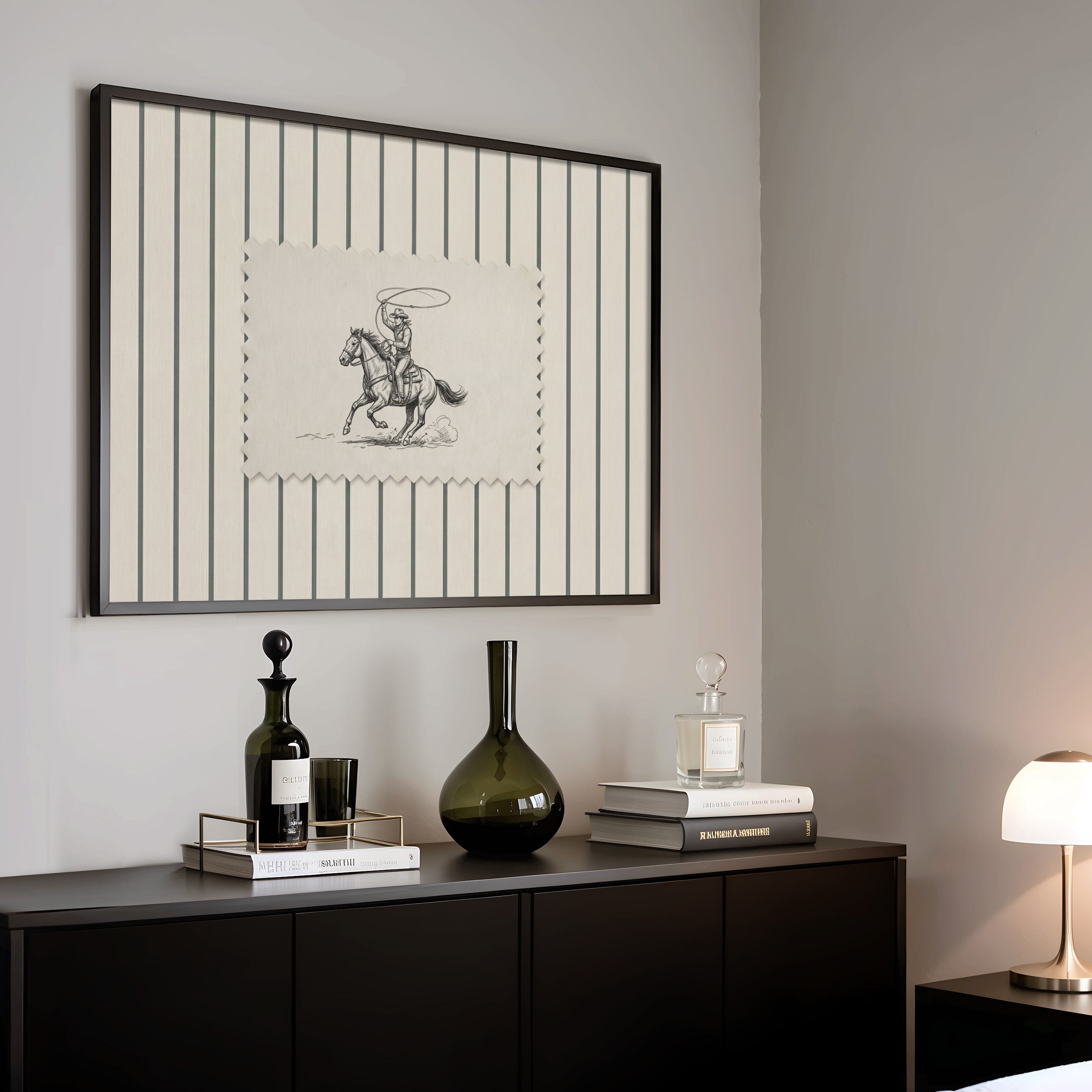 a black and white framed picture of a cowboy on a horse, a black vase, a stack of books, and a lamp on a black dresser.