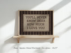a framed sign with the text "YOU'LL NEVER KNOW DEAR HOW MUCH I LOVE YOU" displayed on a wooden shelf against a white wall.