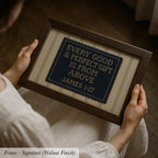 A person is holding a framed cross-stitch embroidery with the text "EVERY GOOD & PERFECT GIFT IS FROM ABOVE JAMES 1:17" on it.