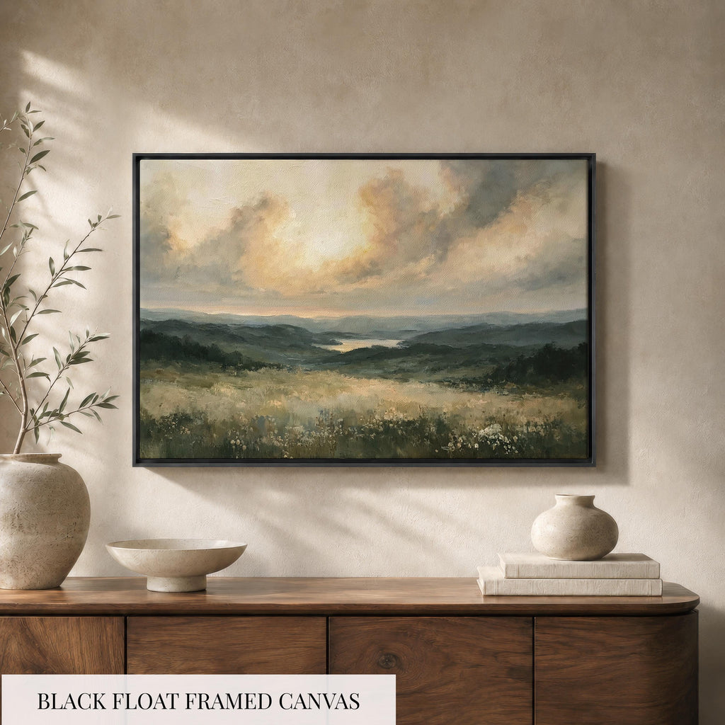 a black float framed canvas depicting a serene landscape with rolling hills, a lake, and a cloudy sky. The painting is hung on a wall above a wooden cabinet, which has a vase and a stack of books on top.