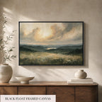 a black float framed canvas depicting a serene landscape with rolling hills, a lake, and a cloudy sky. The painting is hung on a wall above a wooden cabinet, which has a vase and a stack of books on top.