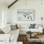 a living room with a large, gold-framed canvas artwork depicting a blue floral design hanging on the wall above a white couch. The room has a rustic, country-style decor with wooden accents and a wooden coffee table.