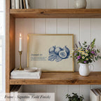 a wooden shelf with a framed print of a blue illustration of fruit, a white vase with flowers, and some books and other decorative items.