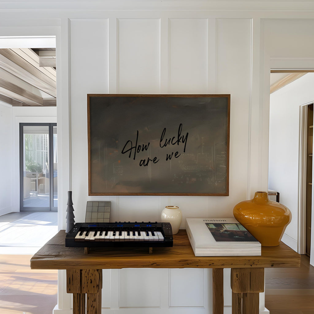 a wooden table with a black keyboard, a white vase, and a book, along with a framed quote that reads "How lucky are we".