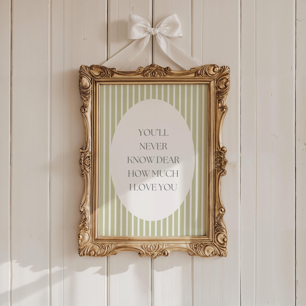 a gold-framed picture with a white border hanging on a white wooden wall. The picture contains a message that reads "YOU'LL NEVER KNOW DEAR HOW MUCH I LOVE YOU".