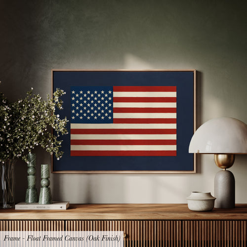 a framed canvas of the American flag hanging on a wall, with a wooden shelf below it and various decorative items such as a lamp, vases, and a book.