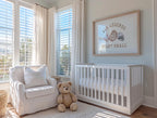 A cozy nursery with a white crib, a white chair, and a teddy bear. The room has large windows that let in natural light, and a framed quote on the wall that says "All legends start small".