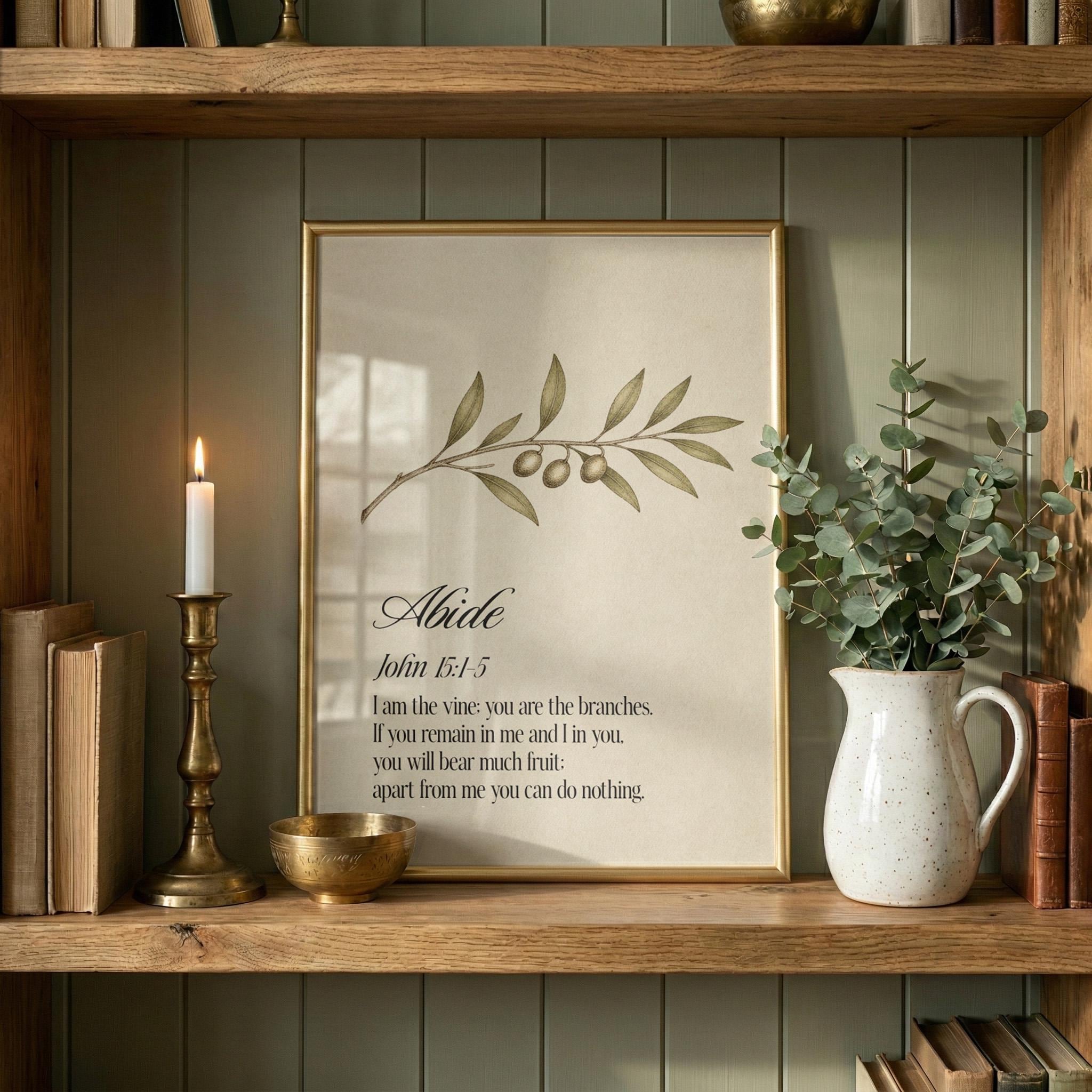 a wooden shelf with a framed print of a biblical verse, a lit candle, a white vase with greenery, and a small bowl.