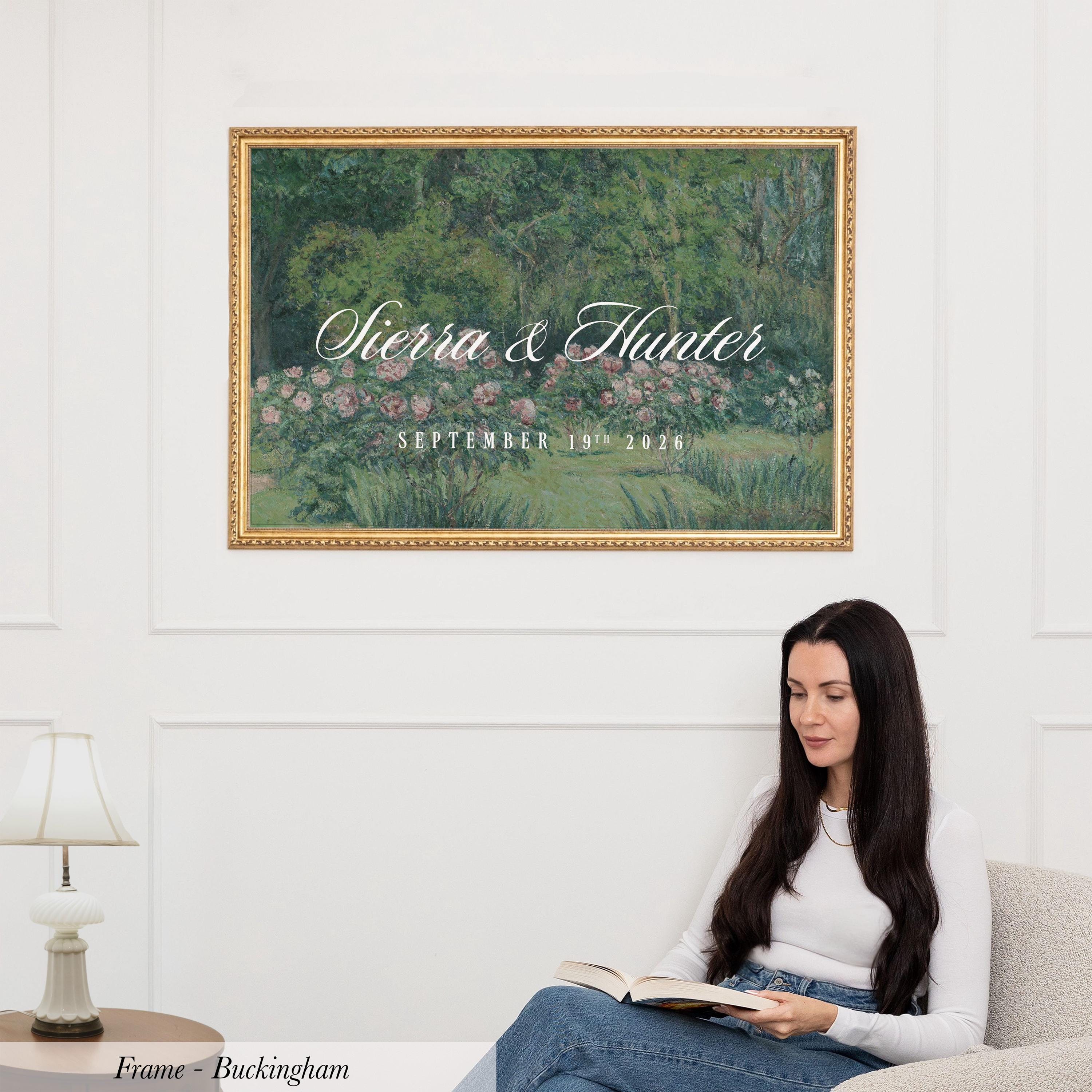 A woman is sitting on a couch, reading a book, with a framed painting of a garden hanging on the wall above her.