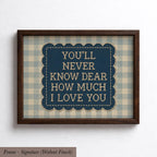 a framed piece of art with a blue background and a scalloped edge. The text within the frame reads "YOU'LL NEVER KNOW DEAR HOW MUCH I LOVE YOU".