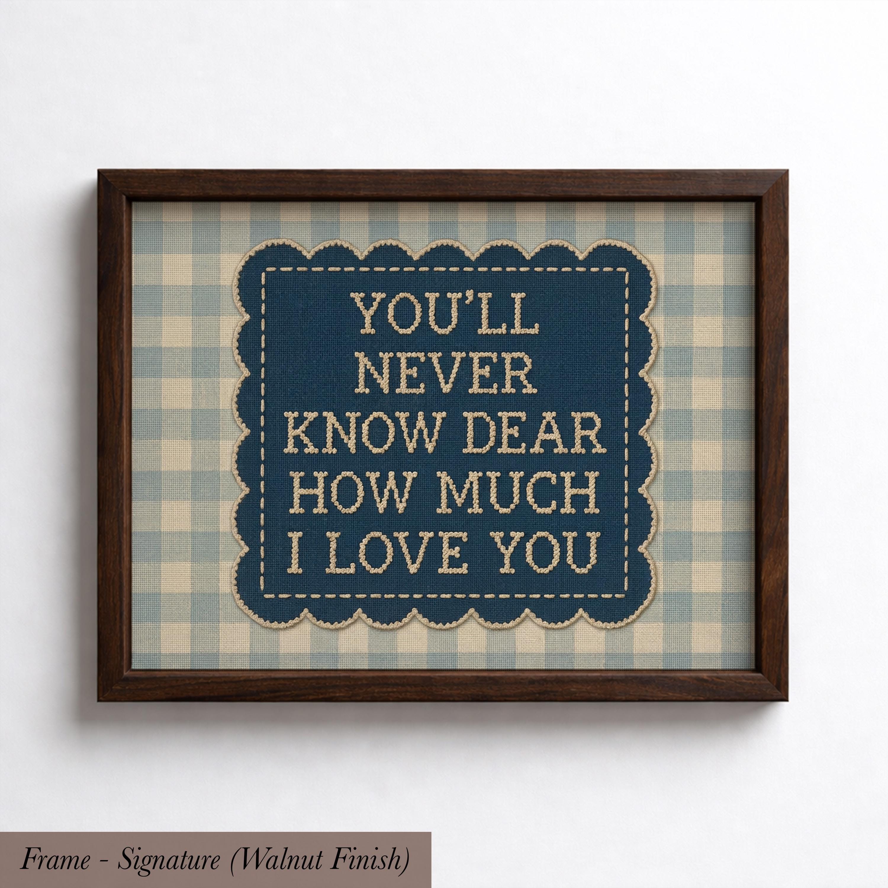 a framed piece of art with a blue background and a scalloped edge. The text within the frame reads "YOU'LL NEVER KNOW DEAR HOW MUCH I LOVE YOU".