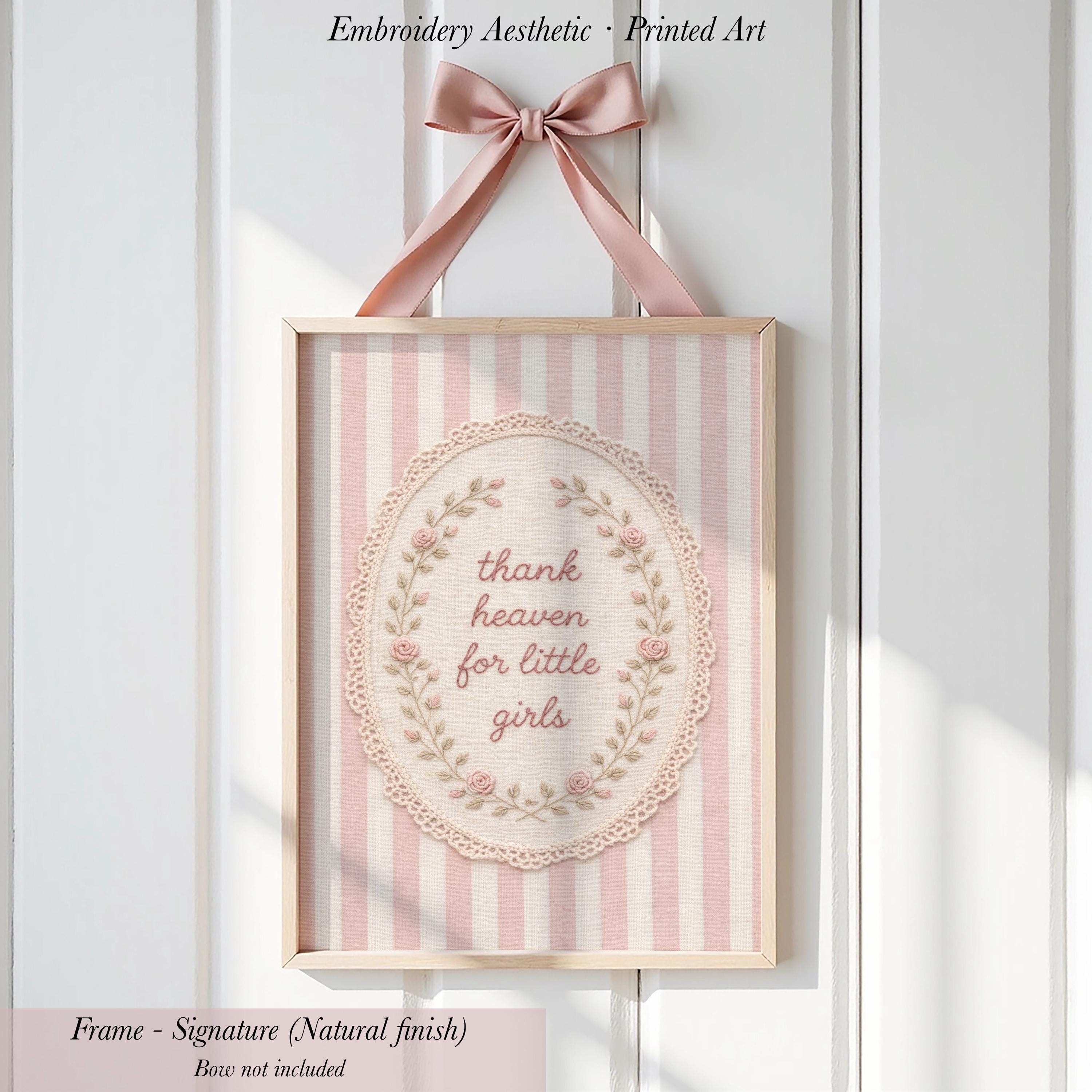 a framed embroidered art piece with a pink and white striped background and a lace border. The text on the art piece reads "Thank heaven for little girls".