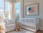 A cozy nursery with a white crib, a white chair, and a teddy bear. The room has large windows that let in natural light, and a framed American flag is displayed on the wall.